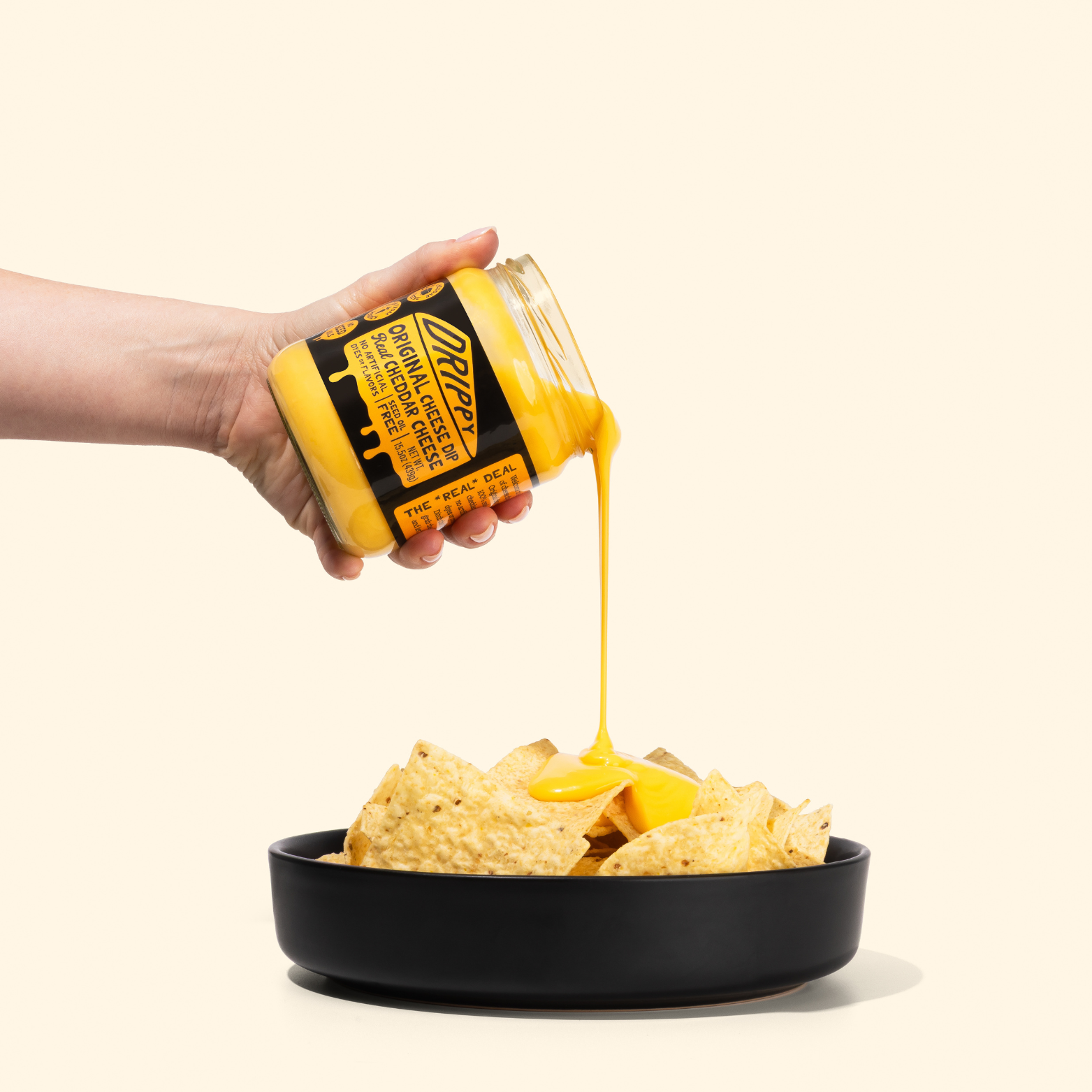 Hand pouring nacho cheese from a jar onto tortilla chips in a black bowl.