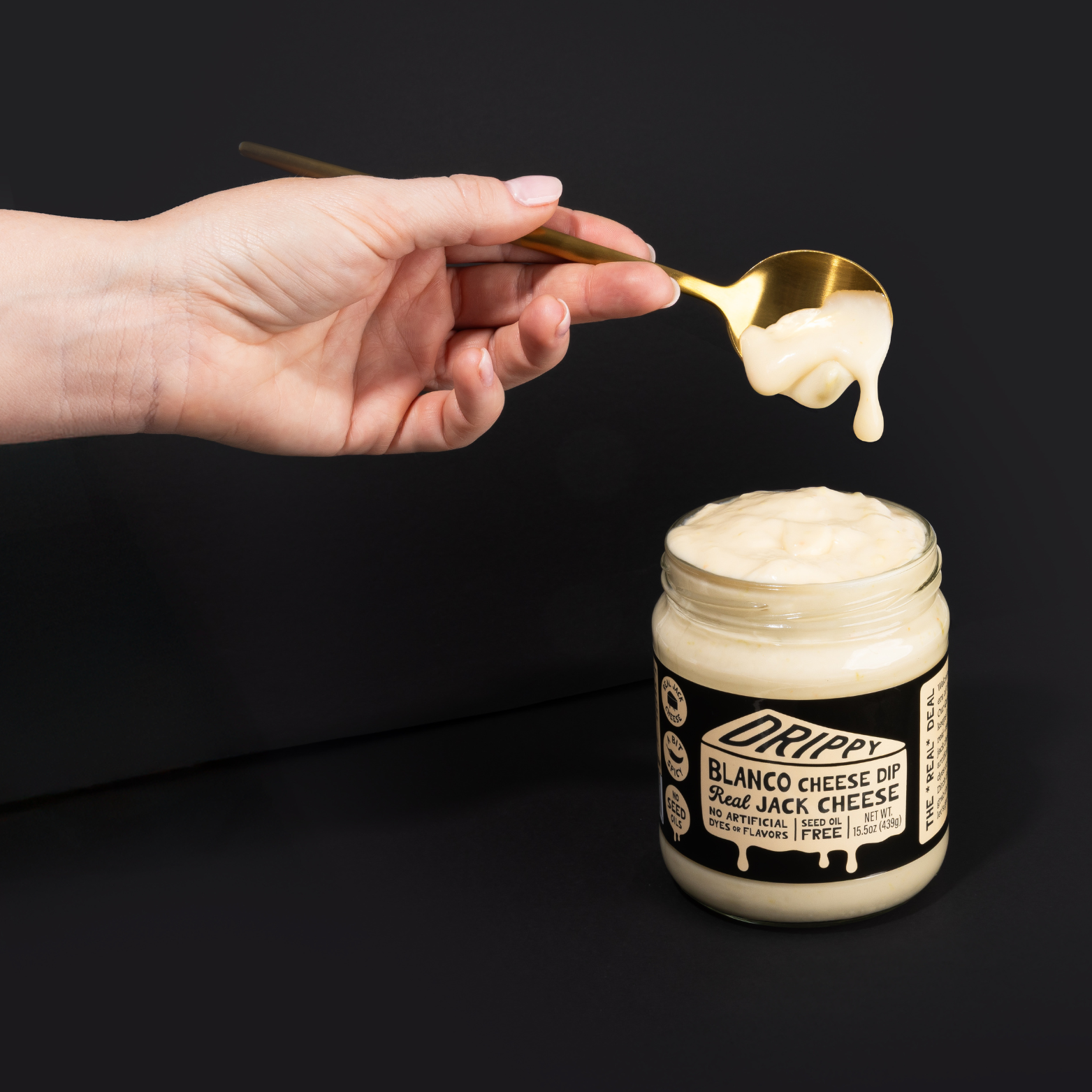 Hand holding a spoon with cheese over a jar of 'Drippy' Blanco Cheese Dip on a black background